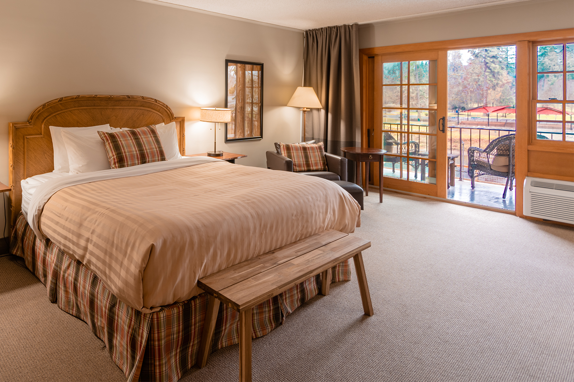 The deluxe king room with a king bed, bench footboard, chair, ottoman side table and wood trimmed sliding glass door to the balcony overlooking the Rogue River and Riverside Park across the river